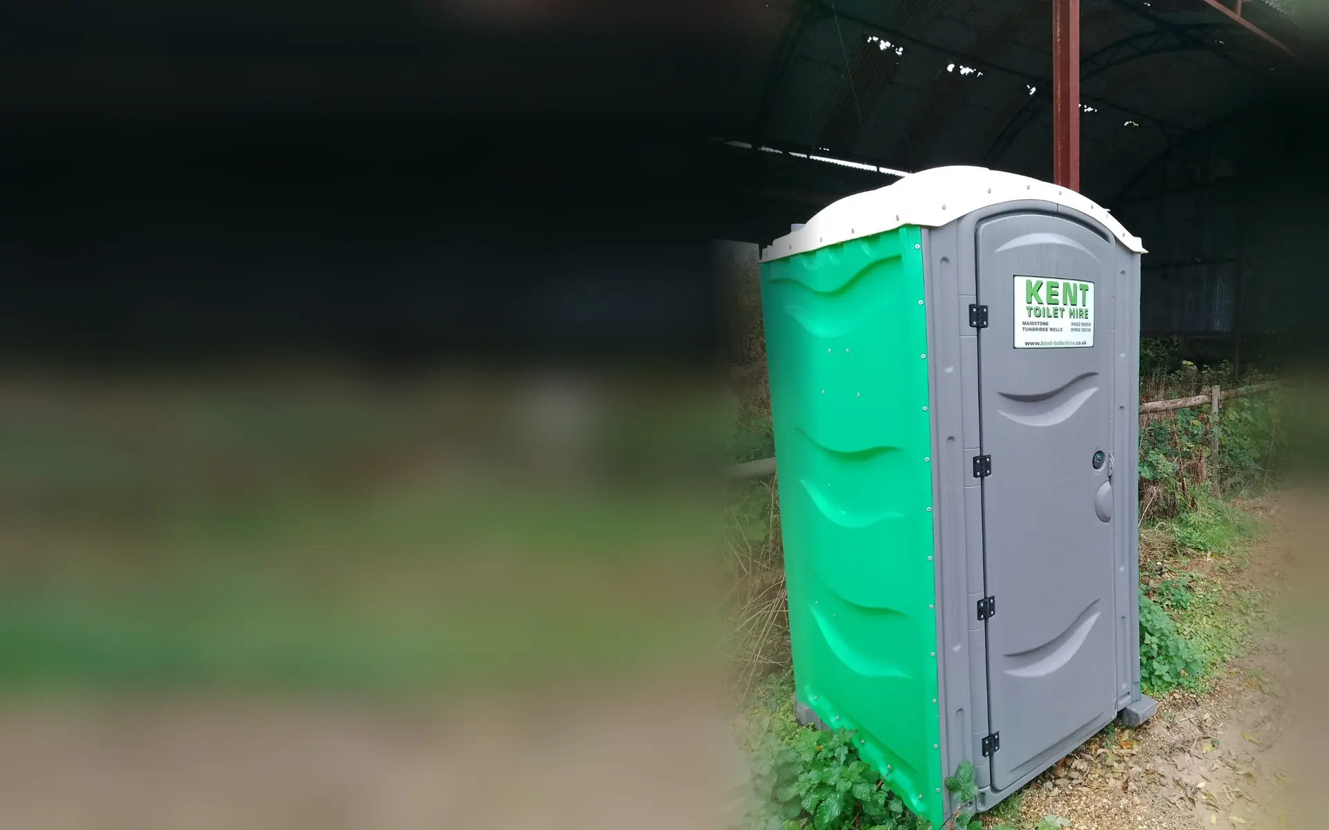 A green and grey Kent Toilet Hire site toilet standing under an open farm barn beside a hedge