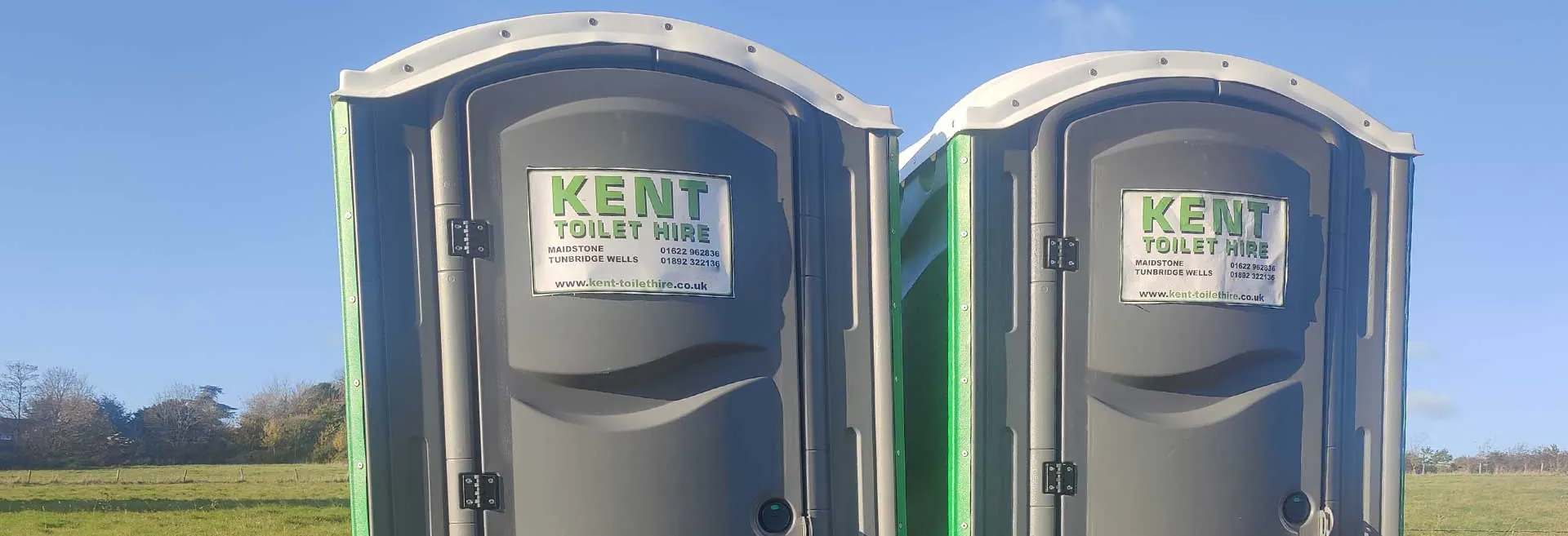 Two of our event toilets with Kent Toilet Hire signs on their doors, in a field by a fence