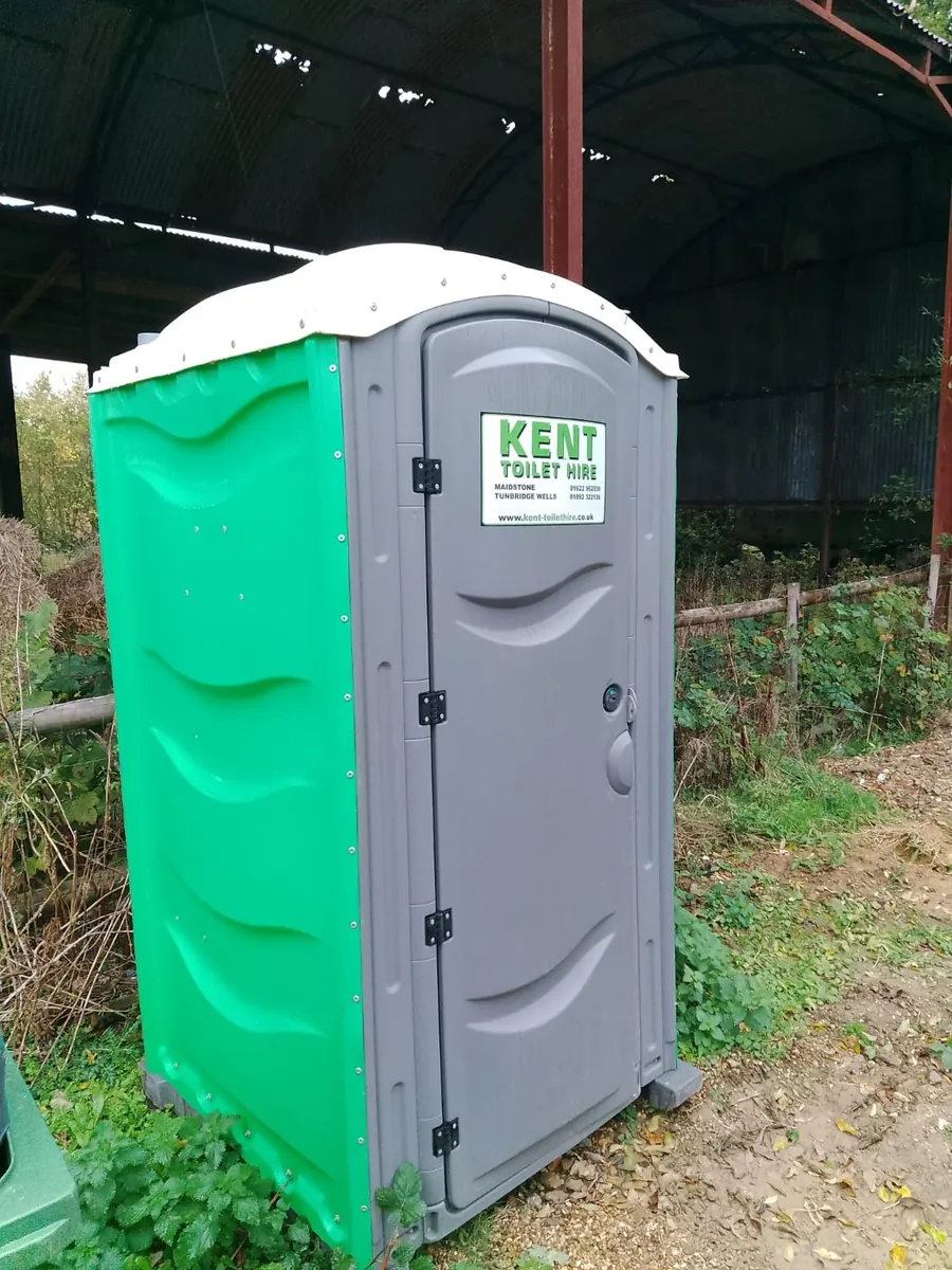 One of our toilets with green sides and the Kent Toilet Hire sign on its door, by an open farm barn