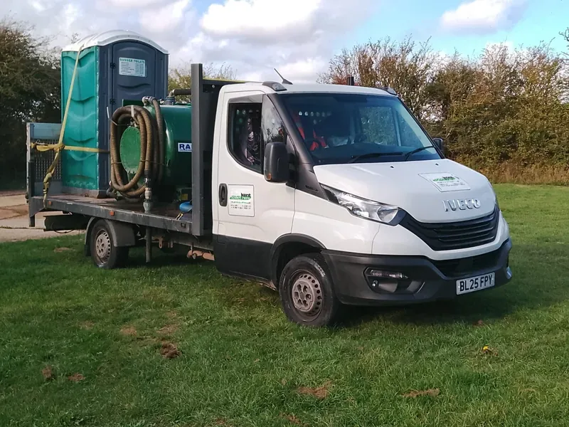 Our white Iveco flatbed truck with a portable toilet and pump hose on the back, parked on grass